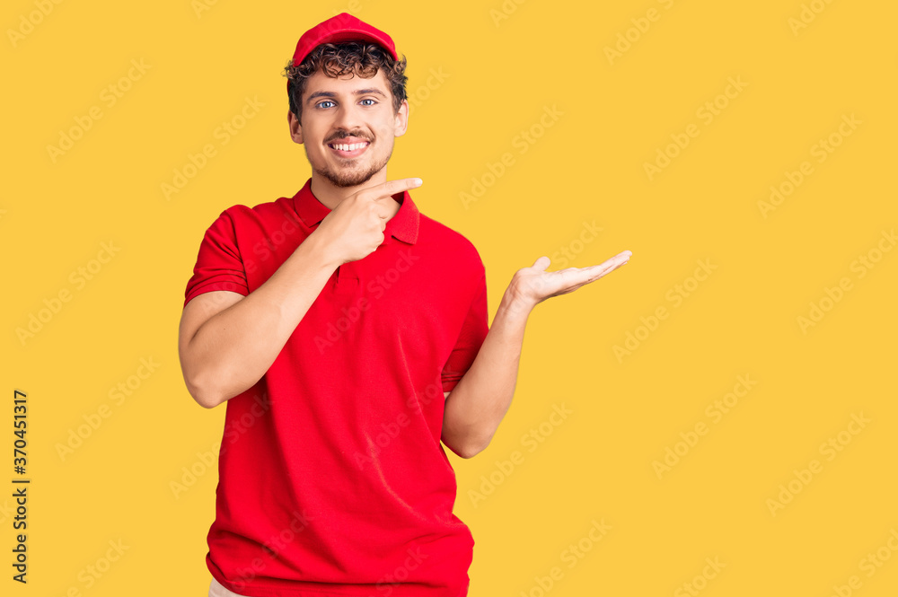 Naklejka premium Young handsome man with curly hair wearing delivery uniform amazed and smiling to the camera while presenting with hand and pointing with finger.