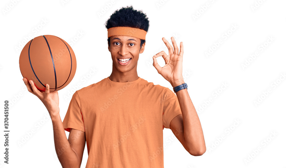Young african american man holding basketball ball doing ok sign with fingers, smiling friendly gesturing excellent symbol