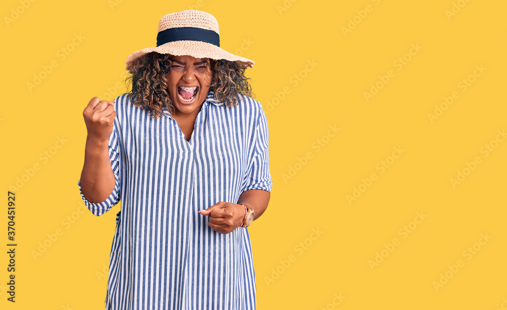 Fototapeta premium Young african american plus size woman wearing summer hat angry and mad raising fist frustrated and furious while shouting with anger. rage and aggressive concept.