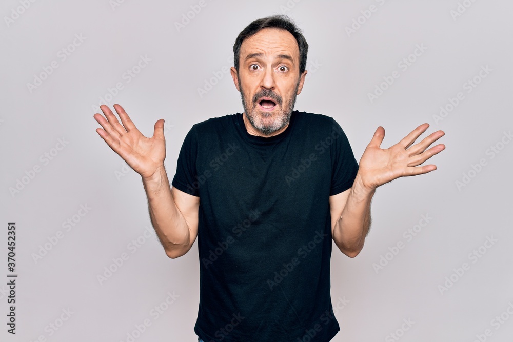 Fototapeta premium Middle age man wearing casual black t-shirt standing over isolated white background clueless and confused with open arms, no idea and doubtful face.