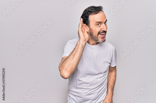 Middle age handsome man wearing casual t-shirt standing over isolated white background smiling with hand over ear listening and hearing to rumor or gossip. Deafness concept.