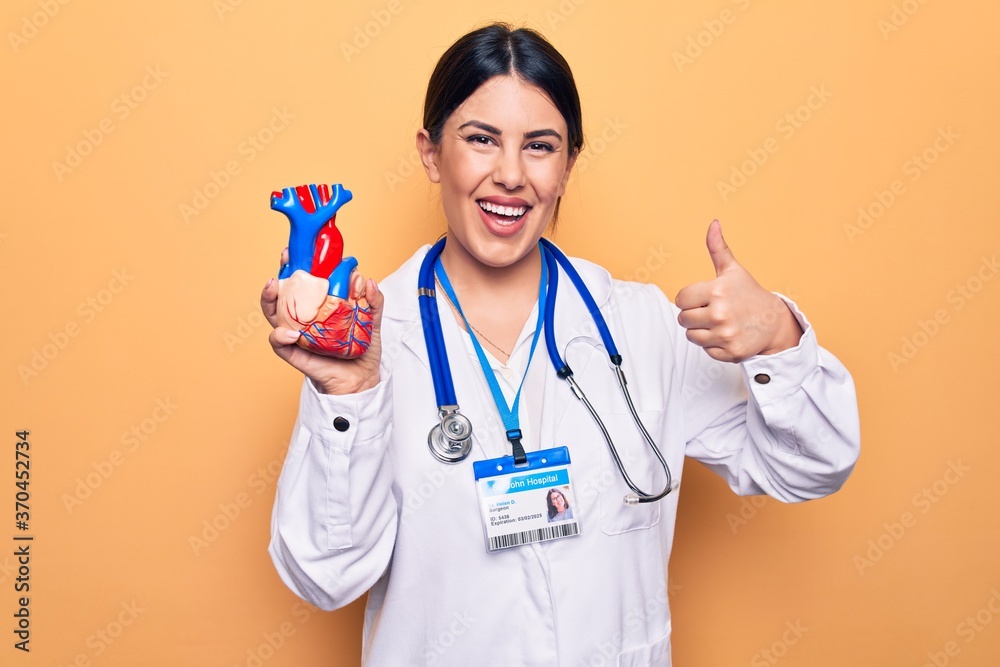 Young beautiful cardiologist woman wearing stethoscope holding heart over yellow background smiling happy and positive, thumb up doing excellent and approval sign Young beautiful cardiologist woman wearing stethoscope holding heart over yellow background smiling happy and positive, thumb up doing excellent and approval sign