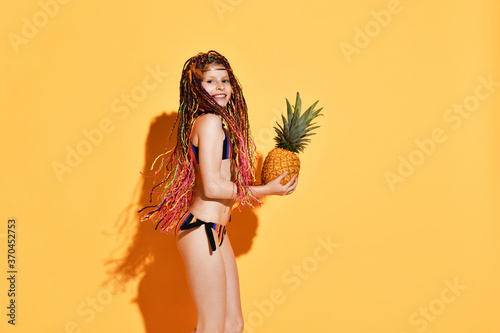Side shot of a girl with colorful afro bunches holding a pineapple. Three quarter shot isolated on orange, copy space.