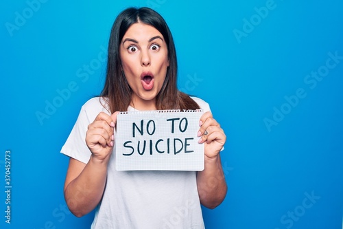 Young beautiful woman asking for psychical problem holding paper with not to suicide message scared and amazed with open mouth for surprise, disbelief face