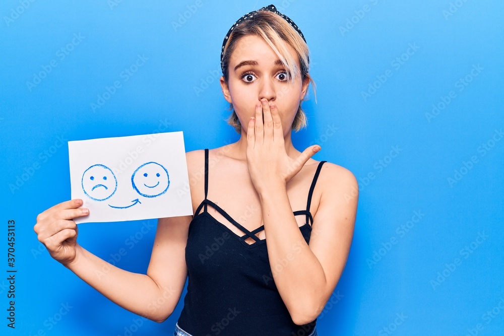 Young beautiful blonde woman holding sad to happy emotion paper covering mouth with hand, shocked and afraid for mistake. surprised expression Young beautiful blonde woman holding sad to happy emotion paper covering mouth with hand, shocked and afraid for mistake. surprised expression