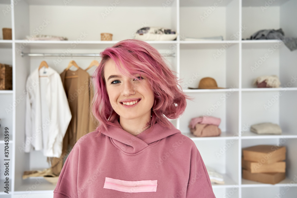 Smiling hipster gen z teen girl fashion designer, blogger with pink hair piercing wearing hoodie looking at camera in front of modern clothes wardrobe closet, face head shot close up portrait. Smiling hipster gen z teen girl fashion designer, blogger with pink hair piercing wearing hoodie looking at camera in front of modern clothes wardrobe closet, face head shot close up portrait.