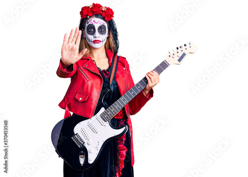 Woman wearing day of the dead costume playing electric guitar with open hand doing stop sign with serious and confident expression, defense gesture