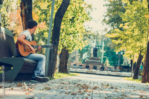 Young man sitting on the bench in the park playing on acoustic guitar with capo. Young attractive man enjoys live music in last sunny days autumn holiday. Retro lens used.