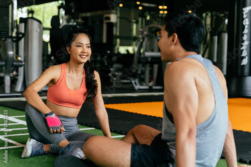 Face of Young Asian mixed sporty woman chatting with a muscuar man sitting on the floor at fitness gym. Healthy couple concept
