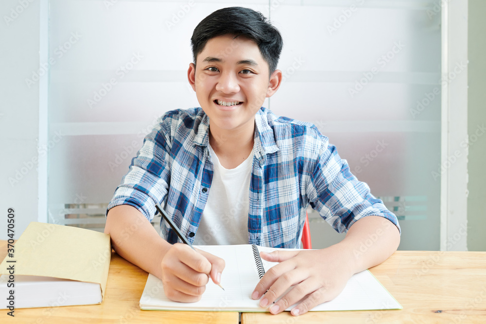 Portrait of smiling Asian schoolboy writing and drawing in copybook when sitting at desk Portrait of smiling Asian schoolboy writing and drawing in copybook when sitting at desk