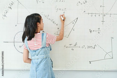 Smart schoolgirl writing on whiteboard when solving geometry task in class, view from the back photo