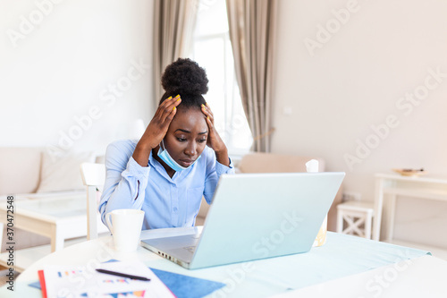 Attractive black young businesswoman sitting alone in her office and coughing as she suffers from a cold. Medical mask and hand disinfectant and stressed woman.