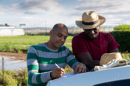 Afro and Latino men farmers signing papers near car on farm