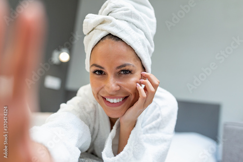 Young beautiful woman with towel on a bed in a hotel room, dressed in a white terry bathrobe, taking selfie on her mobile phone, a slight smile on her face