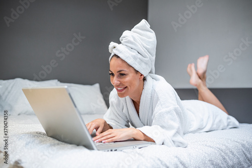 Young beautiful woman with towel on a bed in a hotel room, dressed in a white terry bathrobe, checking her email on her laptop computer, a slight smile on her face
