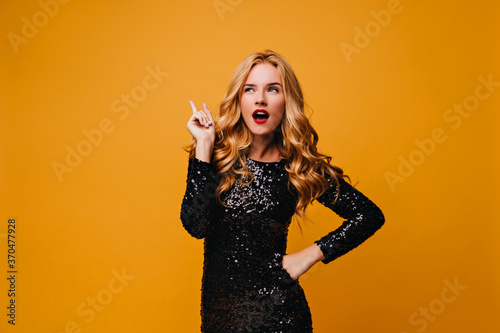 Curious well-dressed girl standing on yellow background. Pensive blonde young woman in black dress thinking about something.