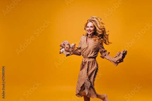 Ecstatic fair-haired woman in vintage dress jumping in studio. Carefree elegant lady in brown outfit dancing on yellow background.
