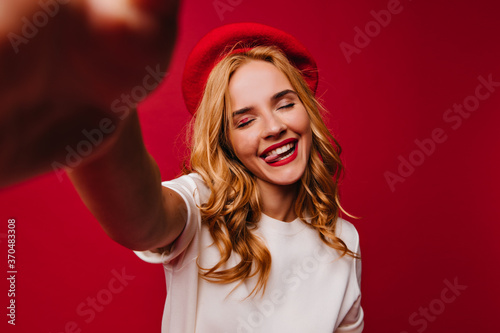 Graceful french girl making selfie on red background with eyes closed. Indoor photo of debonair european woman posing with pleased face expression.