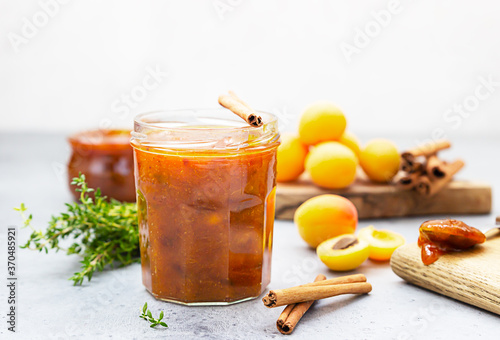 Apricot jam in a glass jar, fresh apricots, thyme and cinnamon on grey stone background.