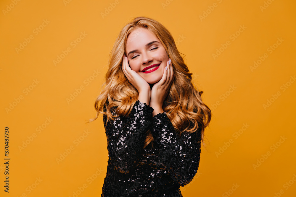 Fototapeta premium Relaxed girl in black dress posing with smile. Studio shot of carefree blonde lady isolated on yellow background.