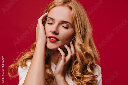 Pleased young woman with blonde hair posing with eyes closed. Carefree fair-haired girl standing on red background.