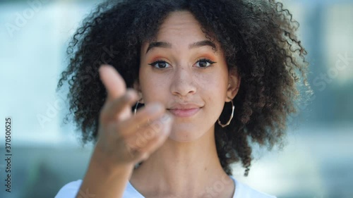 Hey you, come here, join. Beautiful African-American girl, points her index finger at camera, beckons with inviting hand gesture. Young happy woman smiles conspiratorially, intriguingly and playfully.