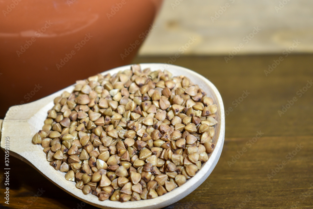 Buckwheat in a wooden spoon, next to a clay pot on a brown wooden background. The concept of healthy dietary food, vegetarianism, traditional Russian cuisine. Selective focus, place for text Buckwheat in a wooden spoon, next to a clay pot on a brown wooden background. The concept of healthy dietary food, vegetarianism, traditional Russian cuisine. Selective focus, place for text