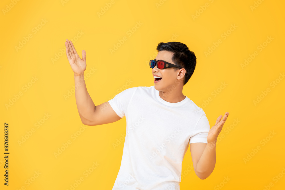 Naklejka premium Fun Portrait of excited young Asian man wearing 3D cinema glasses with both hands raised in isolated studio yellow background