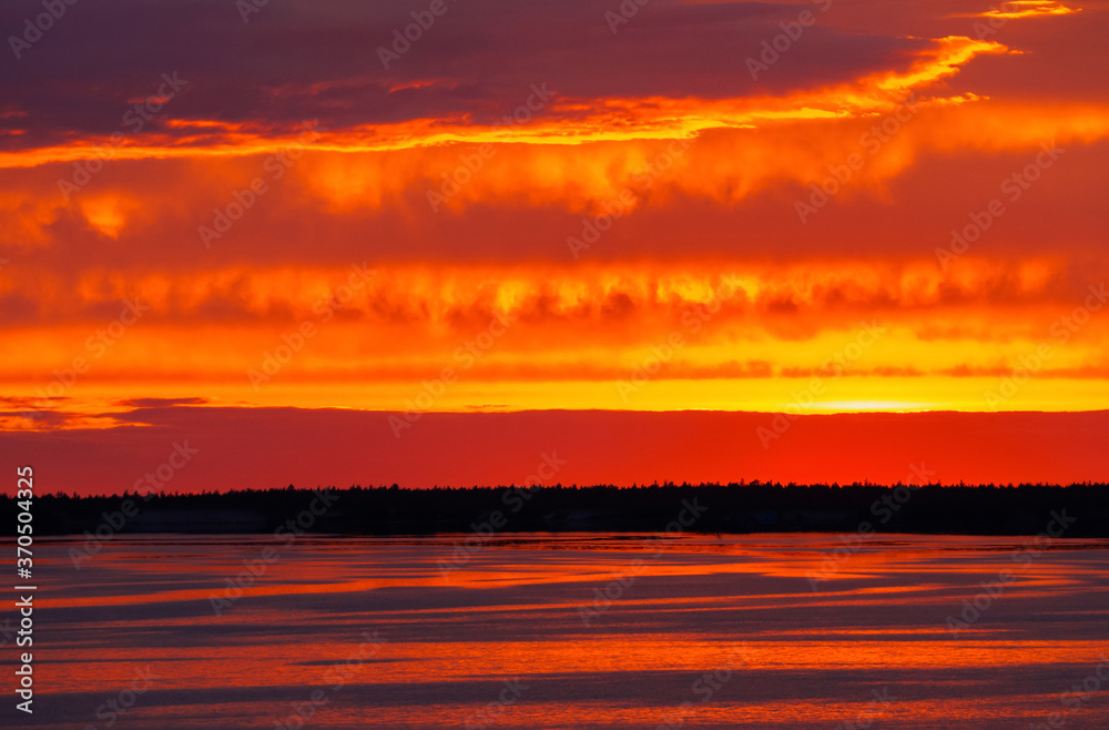 A fiery sunset is reflected in the calm river water, overhanging clouds in the bright orange sky, light waves on the water surface.