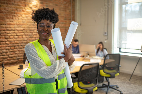 Portrait of young engineer woman with white helmet and blueprints in her hands. Happy female executive with arms crossed. Young businesswoman standing in office.
