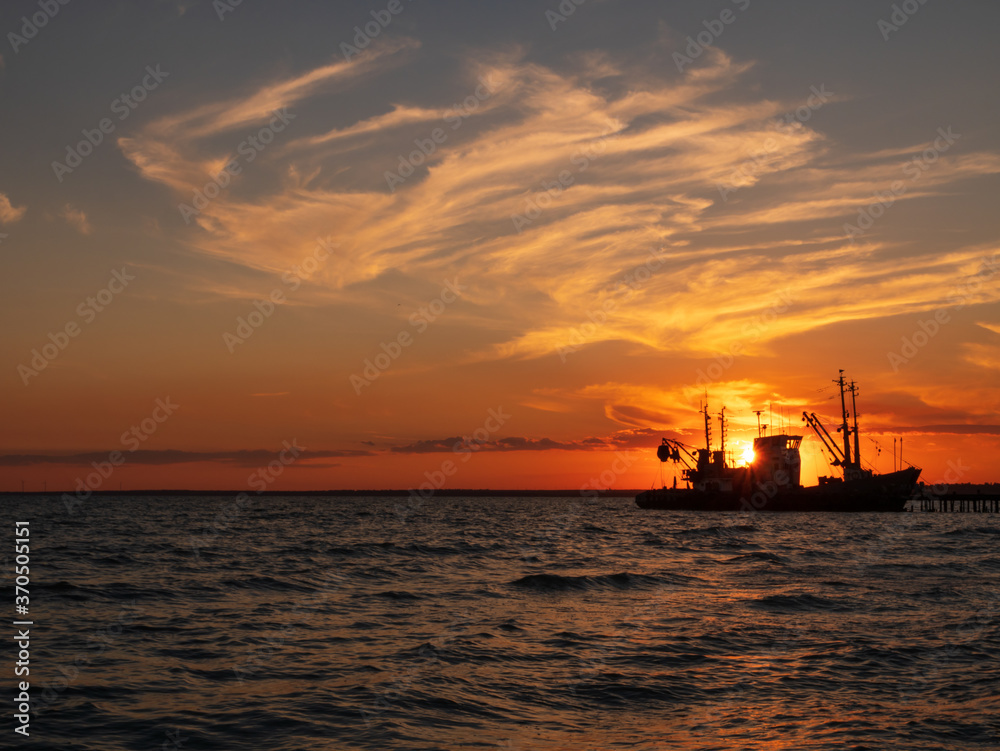 Beautiful orange sunset on sea water. Transport ship on the dock in the evening on seacoast. Shipping logistic by sea. Amazing marine landscape with colorful cloudy sky.