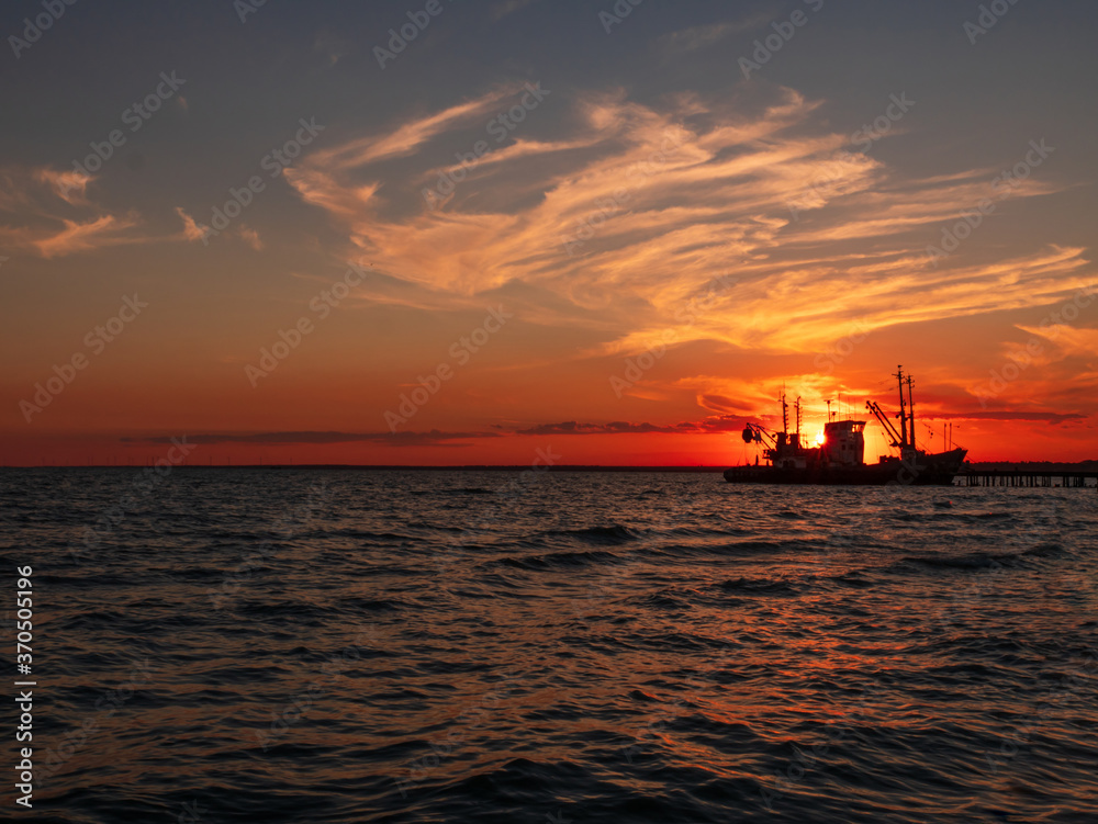 Beautiful orange sunset on sea water. Transport ship on the dock in the evening on seacoast. Shipping logistic by sea. Amazing marine landscape with colorful cloudy sky.