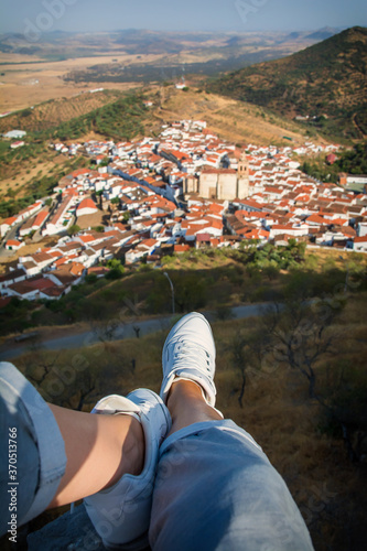 Point of view of a hiker in white sneakers overlooking a picturesque town with white houses in a mountain valley. Hiker relaxing overlooking a city in Extremadura, Spain.