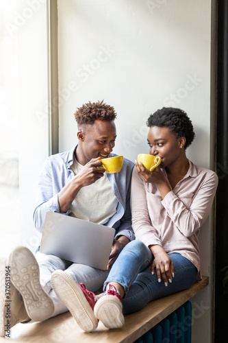 Black man and his girlfriend with laptop drinking coffee while sitting on windowsill at cafe