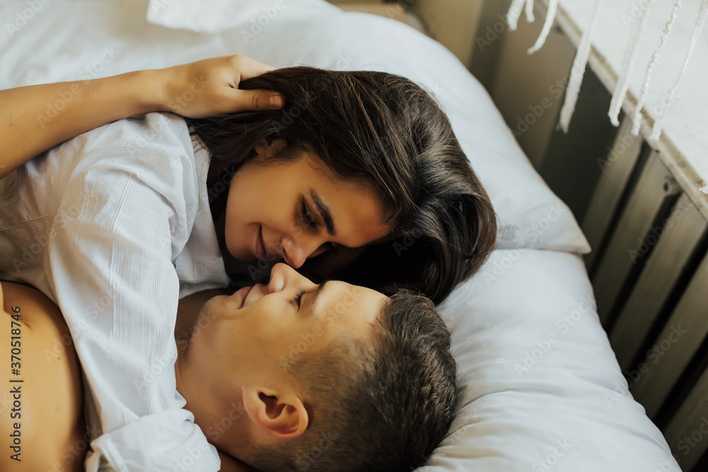 Happy sensual young couple lying in bed together. In love couple hug on the bed near the window in the morning. Love, couple, he and she, together. Side view.