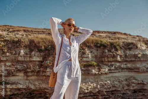 Outdoor summer fashion portrait of elegant woman wearing white linen suit, sunglasses, with round wicker bag, posing on rocks near sea. Copy, empty space for text photo