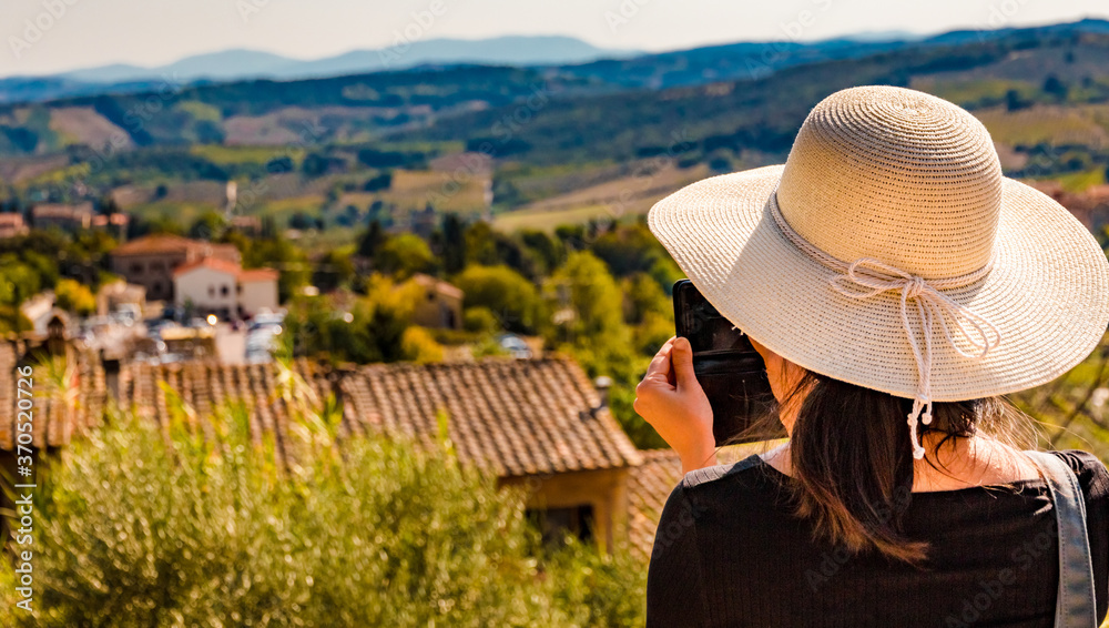 Gorgeous close-up view of a woman, wearing a big sun bonnet and taking photos with her mobile phone of the beautiful countryside of San Gimignano in Tuscany. The picture focuses on the woman! 