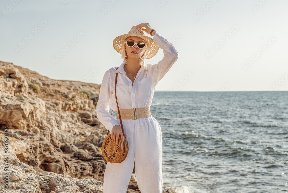 Outdoor summer fashion portrait of elegant woman wearing white linen suit, belt, straw hat, sunglasses, with round wicker bag, posing on rocks near sea. Copy, empty space for text