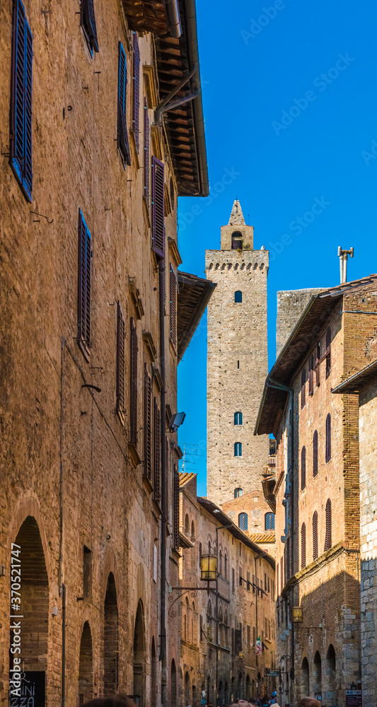 Picturesque portrait shot of the main street Via San Giovanni filled with shops, and the tallest tower Torre Grossa in the background of the famous medieval town San Gimignano, Tuscany, Italy.