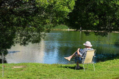 Elderly woman in a white hat sits on a folding chair near a pond in a city park and solves a crossword puzzle on a sunny summer day