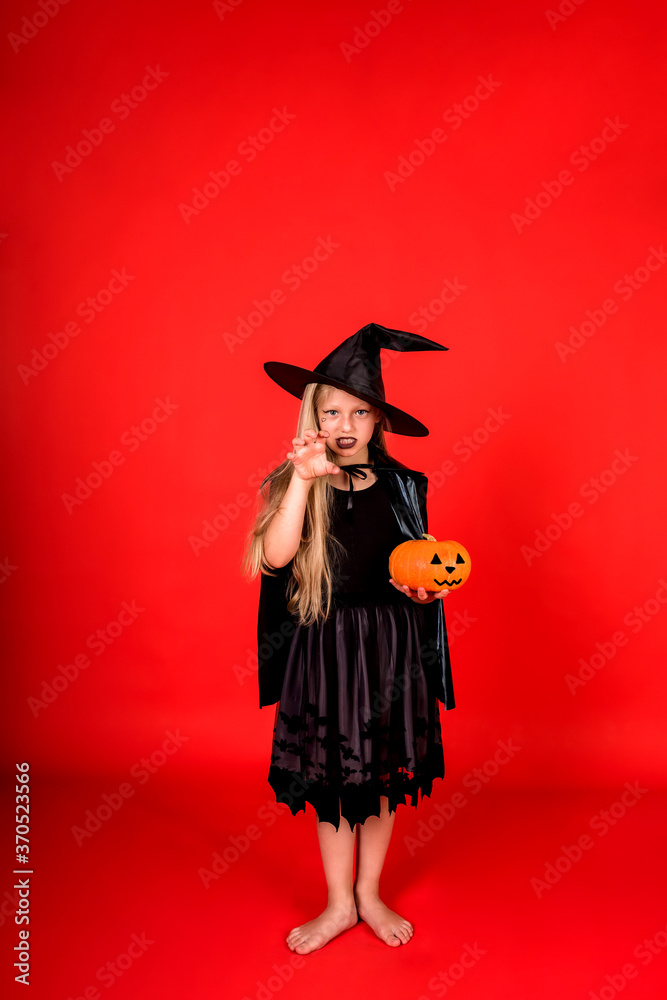 A young girl in a witch costume with a pumpkin hat scares on a red background with a copy of space A young girl in a witch costume with a pumpkin hat scares on a red background with a copy of space