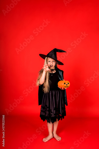 A young girl in a witch costume with a pumpkin hat scares on a red background with a copy of space