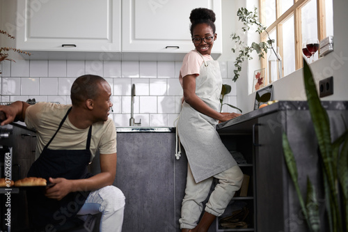 side view on friendly afroamerican married couple in the kitchen, man sit next to oven and talk with wife