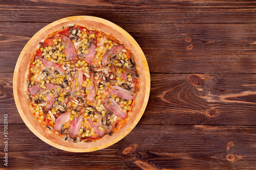 Whole pizza with chicken breast, corn, bacon and mushrooms, on a round wood plate which is on wooden rustic table, top view and copy space