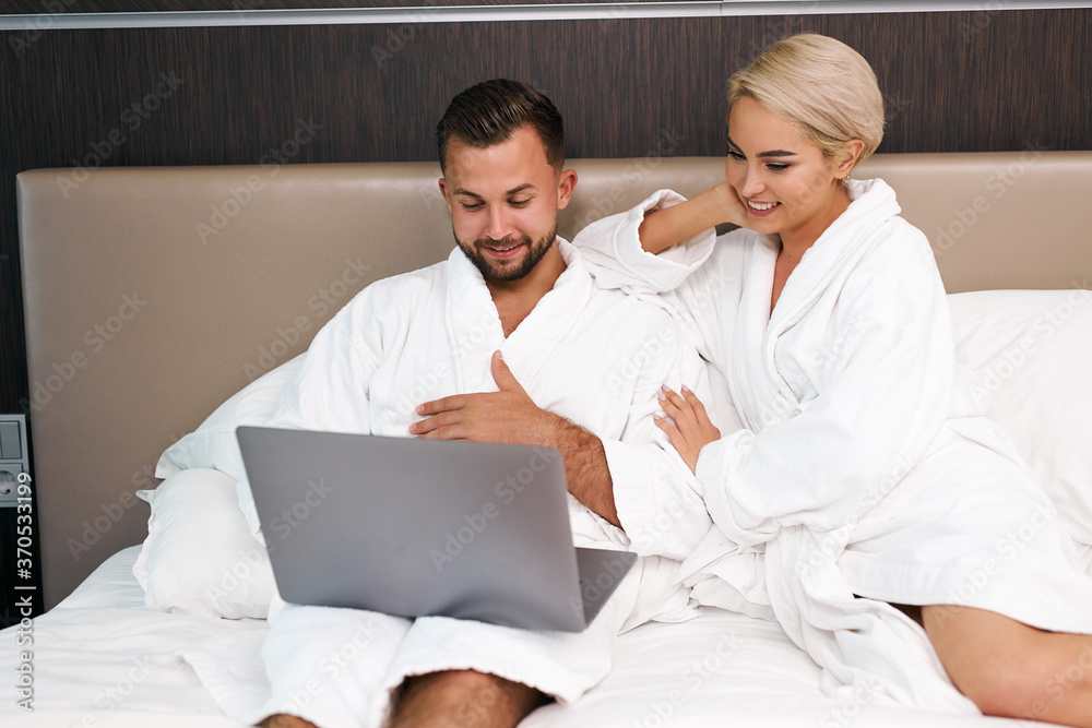 young caucasian couple with laptop in the morning, bearded man and blonde woman discuss where to go during their trip, they lie on bed in bathrobes, in hotel