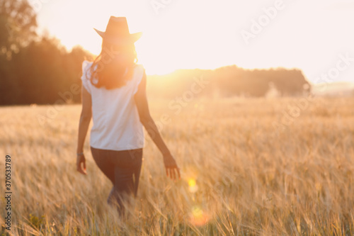 Woman farmer in cowboy hat walking with hands on ears at agricultural wheat field on sunset.