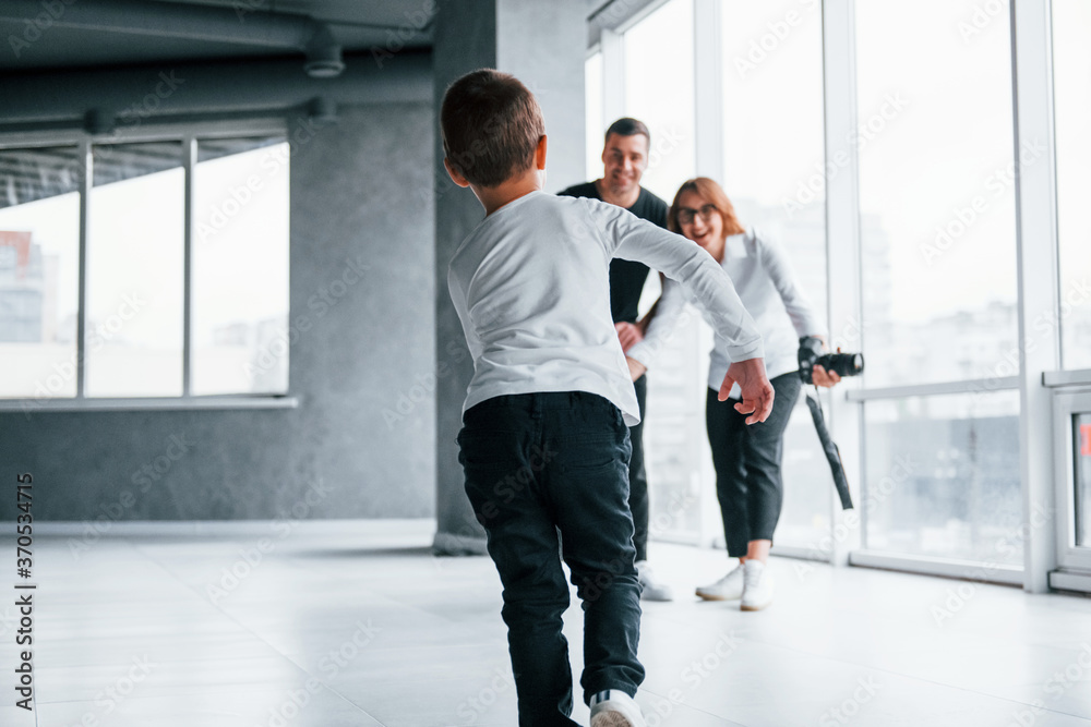 Woman in formal clothes and with camera in hand standing inside of empty room with man and little boy that running and having fun Woman in formal clothes and with camera in hand standing inside of empty room with man and little boy that running and having fun