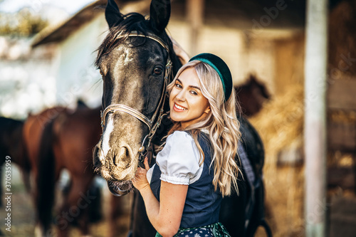 Pretty blonde in traditional dress take care of big black horse at the farm