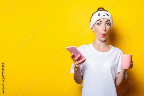 Surprised young girl in a bandage with a phone and a cup of coffee on a yellow background