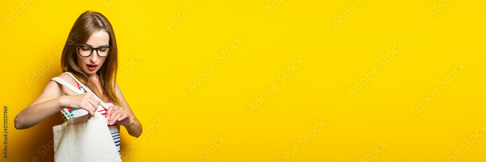 Surprised young woman looking into linen bag with purchases on yellow background. Banner. Surprised young woman looking into linen bag with purchases on yellow background. Banner.